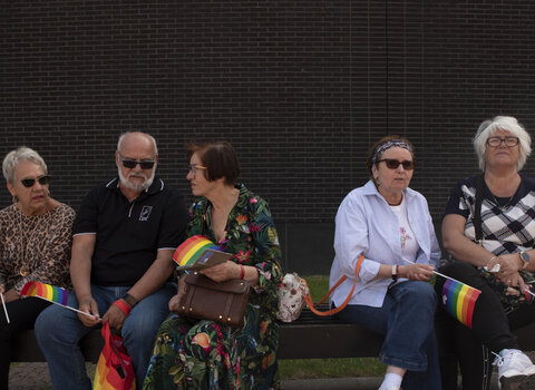 Zdjęcie. Na ławce siedzi pięć osób, cztery kobiety i mężczyzna. Wszyscy są w wieku senioralnym. W dłoniach trzymają tęczowe flagi, a mężczyzna tęczową torbę na zakupy. Siedzą na tle Teatru Szekspirowskiego.