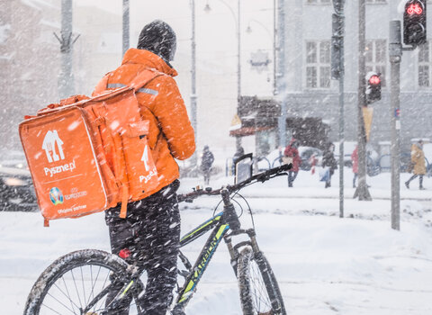 Zdjęcie. Zima, pada gęsto śnieg, pokrył ulice. Na pierwszym planie stoi osoba, kurier na rowerze który przystanął na chwilę. Ma pomarańczową kurtkę i wielki plecak w tym samym kolorze. W głębi widać budynek sądu przy ulicy Nowe Ogrody.