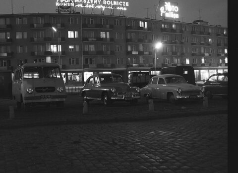 Zabudowa przy ul. Rajskiej w Gdańsku. Zdjęcie czarno-białe. Zdjęcie przedstawia parking samochodowy przed budynkiem. Na nim dwa szyldy: z napisem "Maszyny rolnicze" oraz "Toto Milion" 