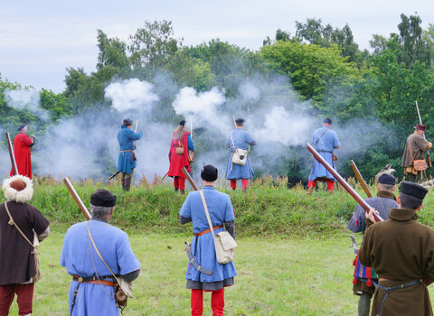 Zdjęcie przedstawia rekonstrukcję walk w Twierdzy Wisłoujście podczas festiwalu rekonstrukcyjnego. Widać jedenaście postaci w historycznych strojach strzelających z muszkietów.