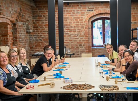 Zdjęcie. Sala edukacyjna Muzeum Bursztynu. Przy dużym stole siedzą uczestnicy warsztatów w fartuchach. Na blacie narzędzia, stos bursztynów, rękawiczki. Uczestnicy są uśmiechnięci i spoglądają w stronę obiektywu.