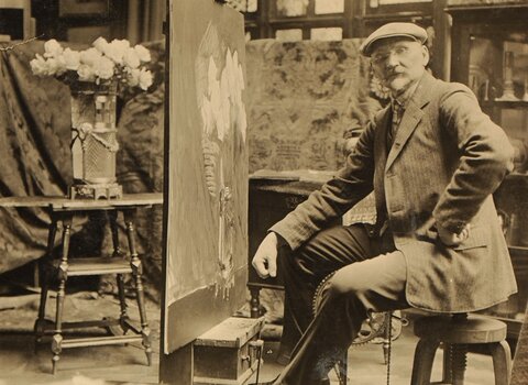 „Leon Wyczółkowski w pracowni”
Czarno-białe zdjęcie przedstawiające malarza siedzącego przy sztalugach w swojej pracowni. Artysta ubrany w garnitur i czapkę, z pędzlem w dłoni, patrzy w stronę aparatu. W tle widoczne są obrazy, tkaniny i wazony z kwiatami, tworzące atmosferę artystycznego skupienia.