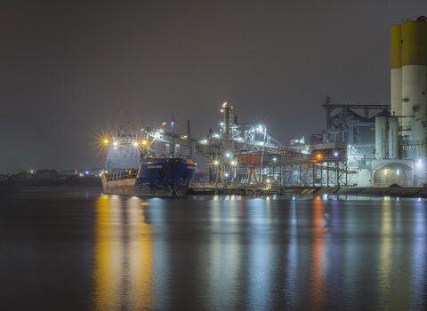 Nocny widok na terminal w Gdańsku. Oświetlone silosy i infrastruktura portowa odbijają się w spokojnej tafli wody, tworząc industrialny pejzaż po zmroku.