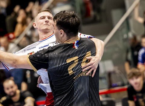 Dwóch zawodników piłki ręcznej w trakcie ostrej walki o piłkę podczas meczu halowego. Jeden z nich oddaje rzut pod presją obrońcy.