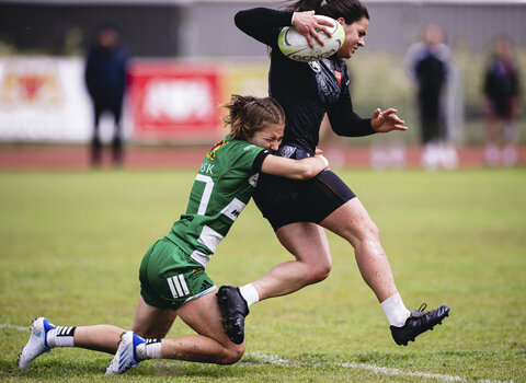 Zawodniczka rugby biegnie z piłką, powstrzymywana przez rywalkę chwytającą ją w pasie. Akcja rozgrywa się na zielonym boisku.