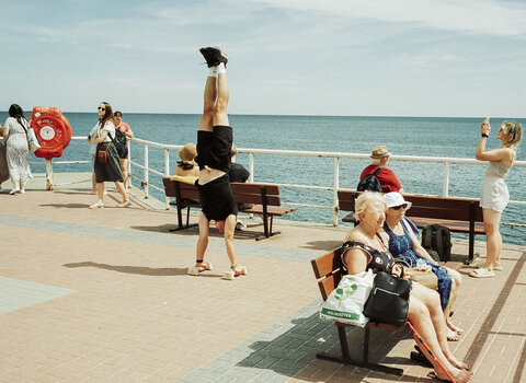 Letnia scena na molo: mężczyzna stoi na rękach, inni spacerują i siedzą na ławkach. W tle spokojne morze i bezchmurne niebo.