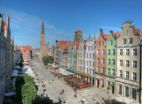 Zdjęcie przedstawia widok na kamienice Długiego Targu na tle błękitnego nieba. W tle wieża Ratusza Głównego Miasta i Dwór Artusa. 