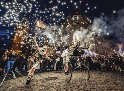 Nocny pokaz uliczny z ogniem: performer w kostiumie rozpala iskry nad głową, obok wózek artystyczny. Wokół zgromadzona publiczność na staromiejskiej ulicy.