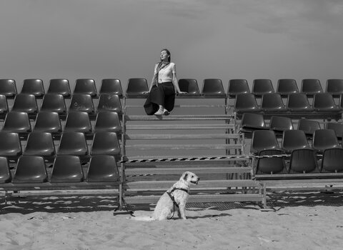 Kobieta siedzi samotnie na środku pustych trybun ustawionych na plaży. Na piasku przed nią siedzi pies; całość w czerni i bieli, z szerokim niebem w tle.