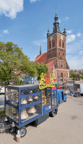 Zdjęcie przedstawia kabinę mobilnego carillonu "Gdańsk". W środku za szybą dzwony. W tle sylwetka kościoła św. Katarzyny.  