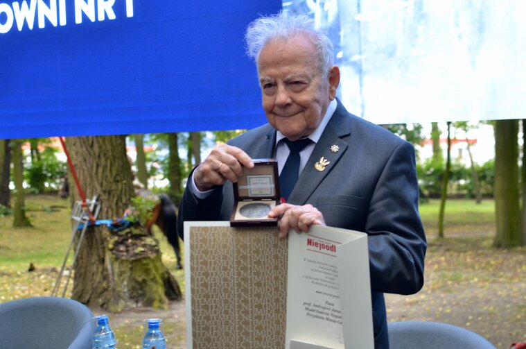 Zdjęcie przedstawia starszego mężczyznę prof. Andrzeja Januszajtisa trzymającego w dłoni dokument oraz medal. W tle fragment ekranu i zieleń.  