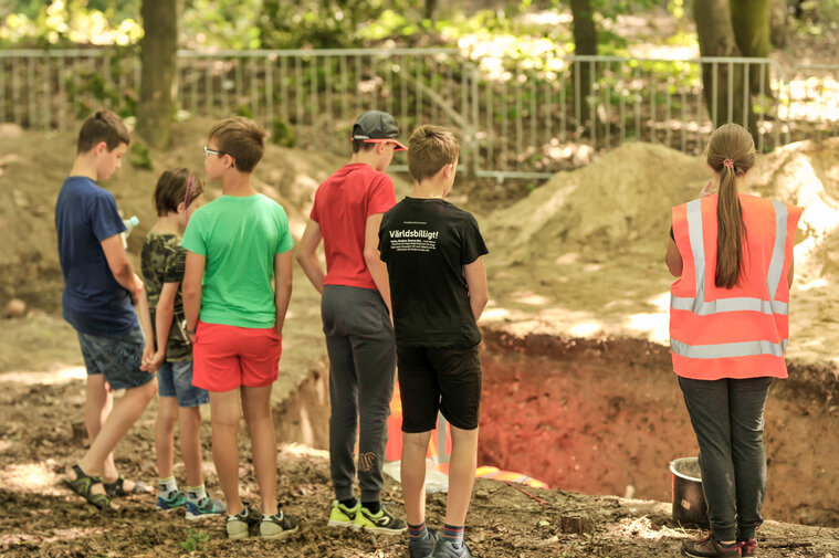 Zdjęcie przedstawia dzieci (po lewej stronie zdjęcia, ukazane od tyłu) patrzące wgłąb wykopu archeologicznego. Po prawej stronie archeolog w kamizelce rozmawiająca z dziećmi. W tle drzewa liściaste. 