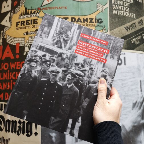 Zdjęcie przedstawia dłoń trzymającą książkę. W tle słup reklamowy z propagandowymi plakatami z lat 30. XX w. w języku niemieckim, na jednym widoczna mapa z konturami II RP z napisem "Polen" i obszarem Wolnego Miasta Gdańska z napisem "Danzig". Na okładce książki w odcieniach szarości widniej Adolf Hitler w otoczeniu niemieckich oficerów. Na okładce czerwone prostokąty z tytułem książki i imieniem oraz nazwiskiem autora.
