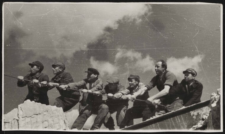 Zdjęcie przedstawia grupę mężczyzn trzymających naprężoną linę na dachu budynku. 