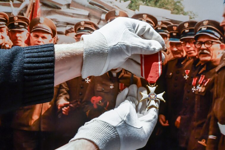 Zdjęcie przedstawia dłonie w rękawiczkach trzymające Order Polonia Restituta. W tle zdjęcie z uroczystości. Widnieją na nim starsi mężczyźni ubrani w mundury. Na piersiach mają odznaczenia. Order trzymany w dłoniach jest podobny do noszonego na mundurze jednego z członków uroczystości, Alfonsa Pillatha (po prawej od dłoni).   