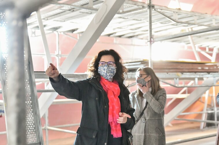 Zdjęcie przedstawia prezydent Gdańska Aleksandrę Dulkiewicz wewnątrz budynku Wielkiego Młyna. Prezydent nosi okulary i maseczkę. Za nią w tle mury z cegły i rusztowania oraz Barbara Frydrych w maseczce. Prezydent trzyma wyciągniętą dłoń w górę wskazując na niewidoczny obiekt. 