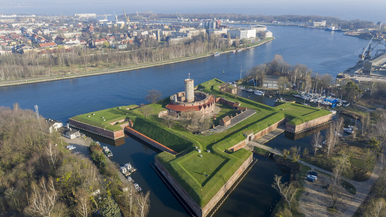 Zdjęcie Twierdzy Wisłoujście wykonane z drona. W centralnym punkcie widoczna wieża oraz otaczające ją kamieniczki oraz bastiony i most wiodący na Twierdzę. W tle ujście Martwej Wisły do Zatoki Gdańskiej. 