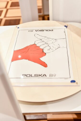 Plakat wyborczy przedstawia dwie dłonie z kciukiem w górę. Dłonie stykają się palcami. Jedna jest biała druga czerwona. Z góry i dołu napis Polska'89