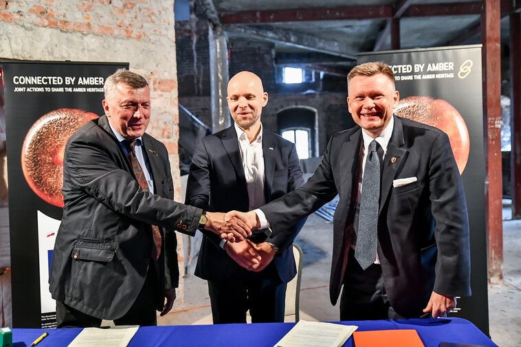 Za stołem od lewej Tadeusz Smolczewski, Alan Aleksandrowicz oraz Waldemar Ossowski ściskają sobie dłonie. Za nimi w tle wnętrza Wielkiego Młyna i surowe ściany oraz dwa postery z bursztynowym paciorkiem. Widok z frontu.