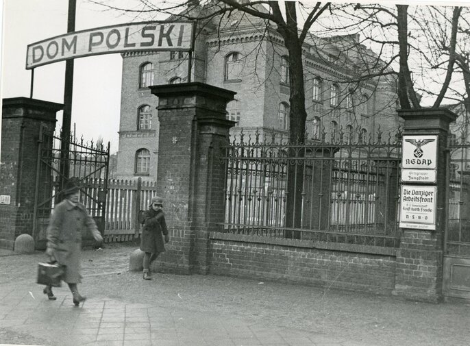 Zdjęcie przedstawia mur a w nim otwartą bramę i w głębi sylwetkę Domu Polskiego przy Wallgasse 16. Przed bramą dwóch przechodniów. Bliżej bramy dziecko w czapce i z plecakiem patrzy w kierunku idącej starszej kobiety w kapeluszu na głowie. Na bramie zawieszone 2 tablice z godłem NSDAP oraz druga z napisem Die Danziger Arbeitsfront.   