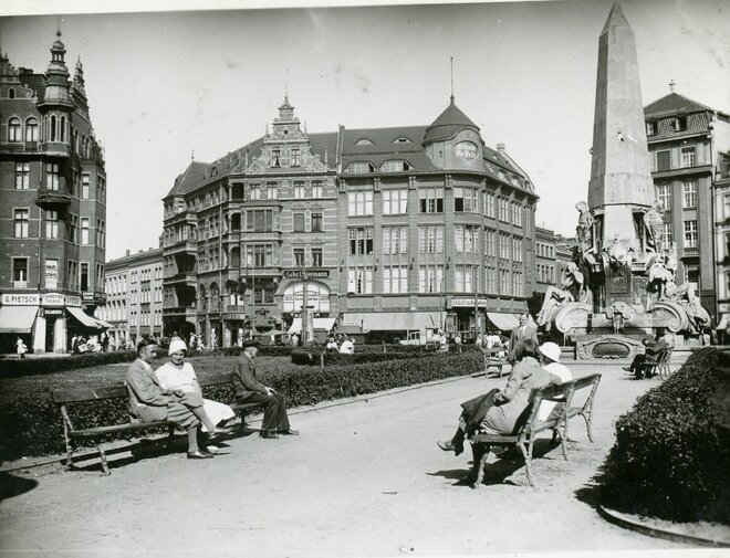 Zdjęcie przedstawia widok na Targ Drzewny. Mężczyźni i kobiety siedzą na ławkach, na prawo od nich obelisk. W centrum zdjęcia fasada budynku.  
