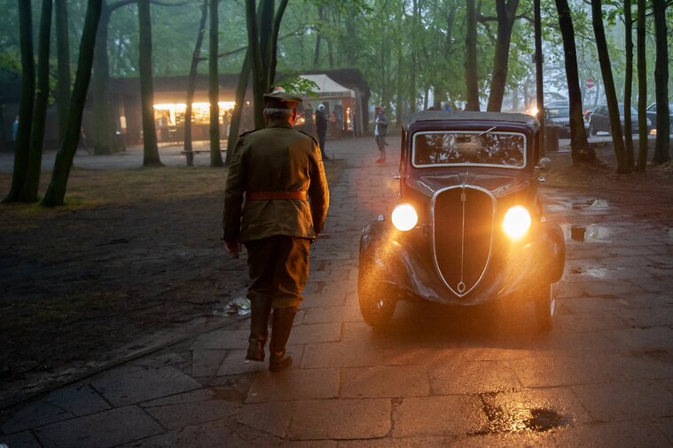 Zdjęcie przedstawia sylwetkę żołnierza Wojska Polskiego z czasów II wojny światowej. W jego kierunku jedzie zabytkowe auto z zapalonymi reflektorami.  