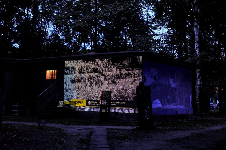 Zdjęcie przedstawia prostopadłościenną bryłę Wartowni nr 1 na Westerplatte. Na jej ścianie frontowej wyświetlany jest czarno-biały film z maszerującymi żołnierzami.  