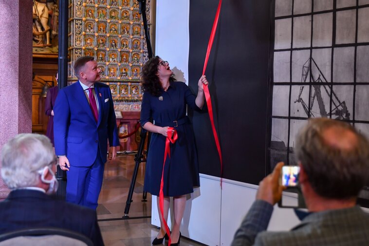 Prezydent Aleksandra Dulkiewicz i Waldemar Ossowski odsłaniają grafikę Dominika Włodarka. Osoby spoglądają się w górę. Prezydent Gdańska trzyma wstęgę. 