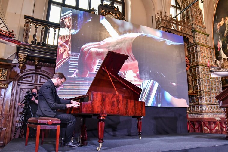 Zdjęcie przedstawia mężczyznę grającego na fortepianie. Klapa instrumentu jest otwarta. Zdjęcie wykonane z tyłu pianisty. 