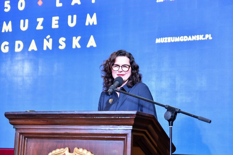 Zdjęcie przedstawia prezydent Gdańska, Aleksandrę Dulkiewicz przemawiającą z mównicy na tle ekrany z napisem 50 lat Muzeum Gdańska.  