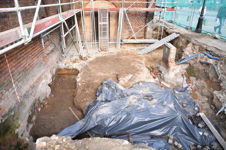 Zdjęcie przedstawia wykop archeologiczny. Od lewej fragmenty muru Wielkiego Młyna, cegłówki i i niżej kamienie. Wokół muru rusztowania. W centrum kadru wkup - betonowa wylewka częściowo przykryta folią. Po prawej stronie fragment płotu.