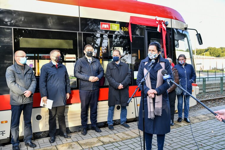 Prezydent Gdańska Aleksandra Dulkiewicz przemawia do uczestników. W głębi, w rzędzie stoją dyrektorzy Muzeum Gdańska oraz zastępca prezydenta Gdańska, Piotr Borawski. W tle sylwetka tramwaju PESA Jazz Duo.  