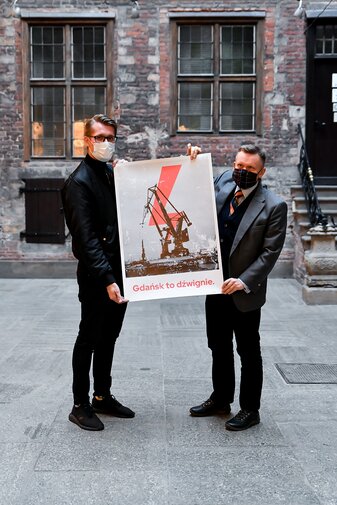 Na zdjęciu Michał Marcinkowski (z lewej) i Waldemar Ossowski (z prawej) trzymają w dłoniach plakat. Na plakacie żuraw portowy, piorun i poniżej hasło „Gdańsk to dźwignie”. W tle fragment nowożytnych murów wewnątrz dziedzińca Ratusza Głównego Miasta.  

Kadr w pionie.