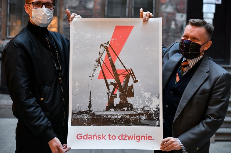 Na zdjęciu Michał Marcinkowski (z lewej) i Waldemar Ossowski (z prawej) trzymają w dłoniach plakat. Na plakacie żuraw portowy, piorun i poniżej hasło „Gdańsk to dźwignie”. W tle fragment nowożytnych murów wewnątrz dziedzińca Ratusza Głównego Miasta.  