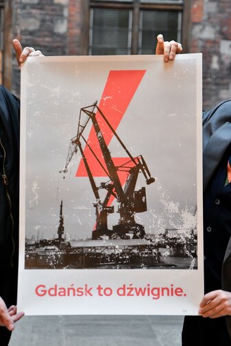 Przekazany przez Michała Marcinkowskiego plakat. Na nim żuraw portowy zakrywający fragmenty dominującego nad nim pioruna. Poniżej hasło „Gdańsk to dźwignie”. Plakat trzymają dwie pary dłoni.   

