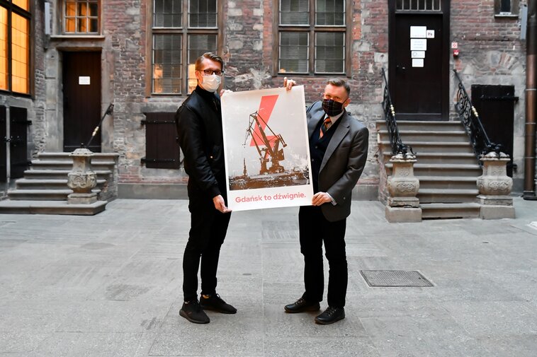 Na zdjęciu Michał Marcinkowski (z lewej) i Waldemar Ossowski (z prawej) trzymają w dłoniach plakat. Na plakacie żuraw portowy, piorun i poniżej hasło „Gdańsk to dźwignie”. W tle fragment nowożytnych murów wewnątrz dziedzińca Ratusza Głównego Miasta.  

Kadr w poziomie.