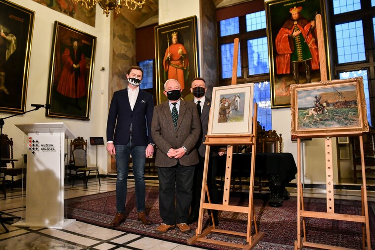 Na zdjęciu od lewej: Zastępca Prezydent Gdańska Piotr Grzelak, Bogdan Radys oraz Waldemar Ossowski stoją w Wielkiej Sali Wety Ratusza Głównego Miasta. Po prawej dwie sztalugi i dwa obrazy Wojciecha Kossaka. 