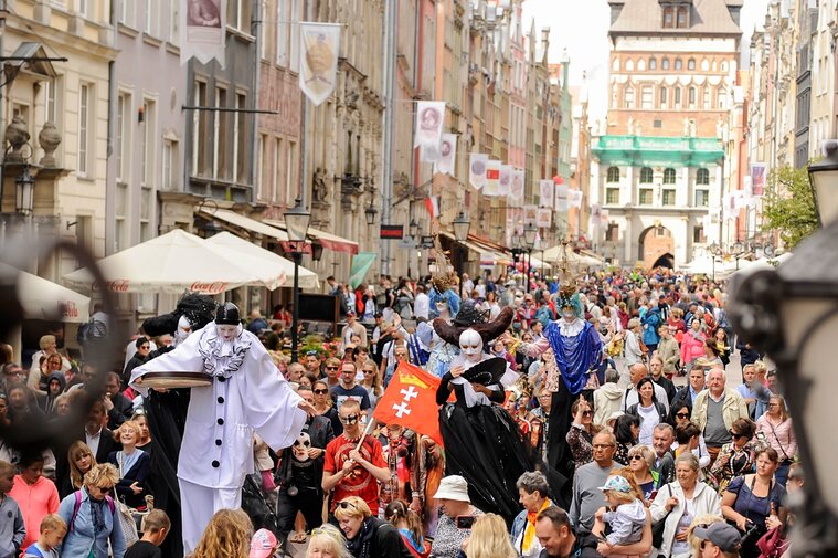 Parada – Gdańsk  Zdjęcie przedstawia paradę na ulicy Długiej w Gdańsku. Szczudlarze oraz modele i modelki w strojach karnawałowych i maskach wzorowanych na strojach weneckich. W paradzie uczestniczą również liczni mieszkańcy. Fot. Agnieszka Grabowska