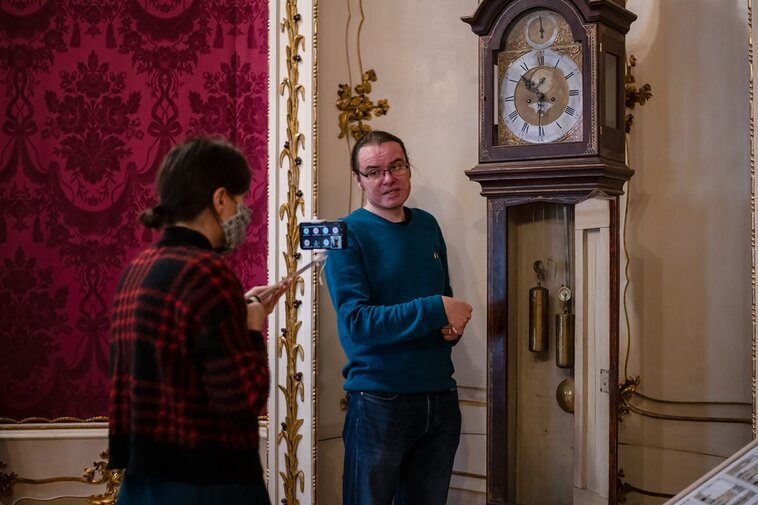 Zdjęcie przedstawia dwie osoby. Kobieta trzyma kijek, a na nim telefon. Po lewej w głębi długowłosy mężczyzna w okularach patrzy w obiektyw i opowiada do kamery o starym zabytkowym zegarze. Fot. A. Grabowska (Muzeum Gdańska)