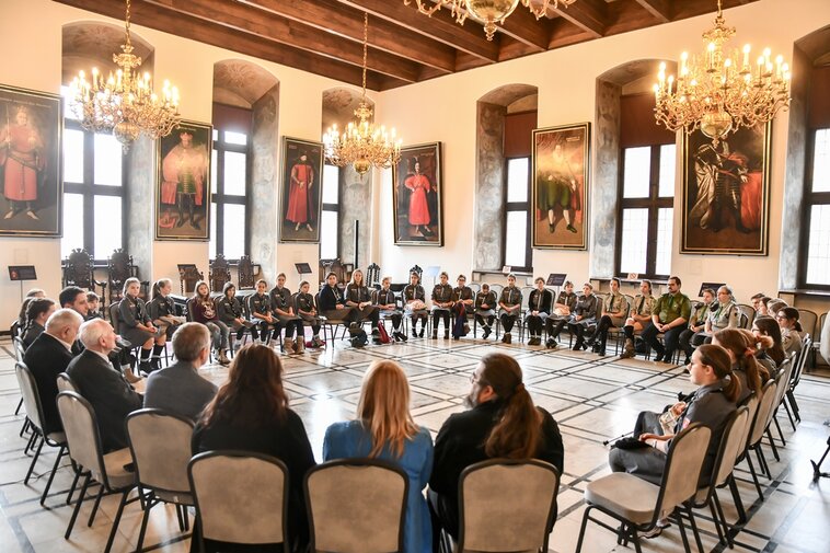 Zdjęcie kolorowe. Wewnątrz dużej sali na ustawionych w okręgu krzesłach siedzą harcerze i kombatanci. Na ścianach sali wiszą duże portrety królów Polski. Z sufitów zwisają ozdobne żyrandole.