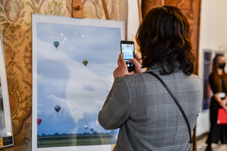 Zdjęcie. Kobieta stojąca plecami do widza fotografuje telefonem komórkowym fotografię przedstawiającą wzlatujące w zachmurzone niebo balony. 