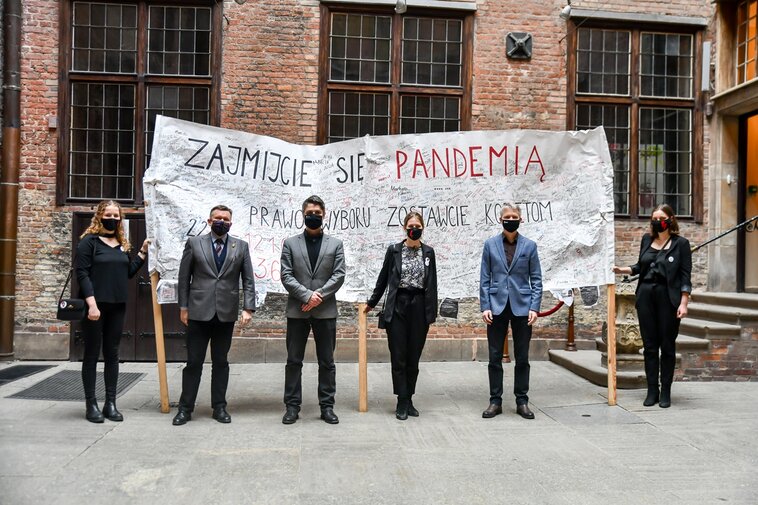 Zdjęcie. Dziedziniec. Od lewej Julia Landowska, Waldemar Ossowski, Andrzej Gierszewski, Aleksandra Łoboda, Janusz Marszalec i Marianna Łoboda. Za nimi transparent z napisem "Zajmijcie się pandemią".