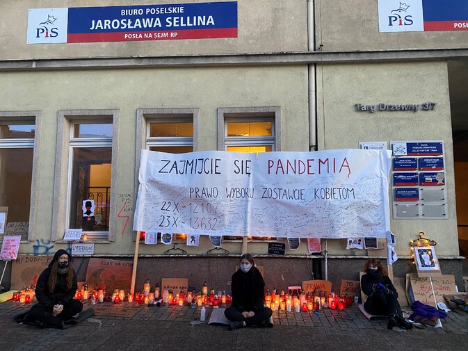 Zdjęcie. Fasada budynku. Transparent. Pod nim w otoczeniu zniczy, siedzą trzy kobiety w maseczkach.  
