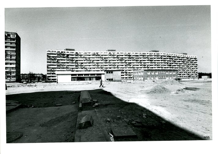 Przykład fotografii poszukiwanych przez Muzeum Gdańska w projekcie Historie Gdańskich Dzielnic. Czarno-biała fotografia. Plac budowy. Na horyzoncie z prawej mniejszy budynek, za nim falowiec. Pośrodku zdjęcia spacerujący mężczyzna. 