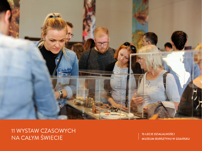 Grupa osób ogląda wystawę. W gablotach leżą eksponaty z bursztynu. Napis 11 wystaw czasowych na całym świecie. 15-lecie działalności Muzeum Bursztynu w Gdańsku.