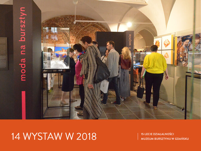 Wnętrze Galerii Palowej, wystawa bursztynu. Pomiędzy gablotami stoi grupa zwiedzających, rozmawiają. Napis 14 wystaw w 2018. 15-lecie działalności Muzeum Bursztynu w Gdańsku.