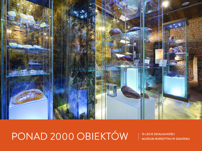 Gabloty wystawowe z naturalnymi bryłami bursztynu. W szklanych ścianach odbija się światło i wnętrze pomieszczenia. Napis Ponad 2000 obiektów. 15-lecie działalności Muzeum Bursztynu w Gdańsku.
