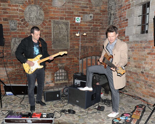 Koncert na dziedzińcu Muzeum Bursztynu. Po lewej Tymon Tymański, gra na gitarze z żółtego bursztynu. Po prawej Wojciech Mazolewski, gra na zwyczajnej gitarze. Obok leżą kable, wzmacniacz, futerał.