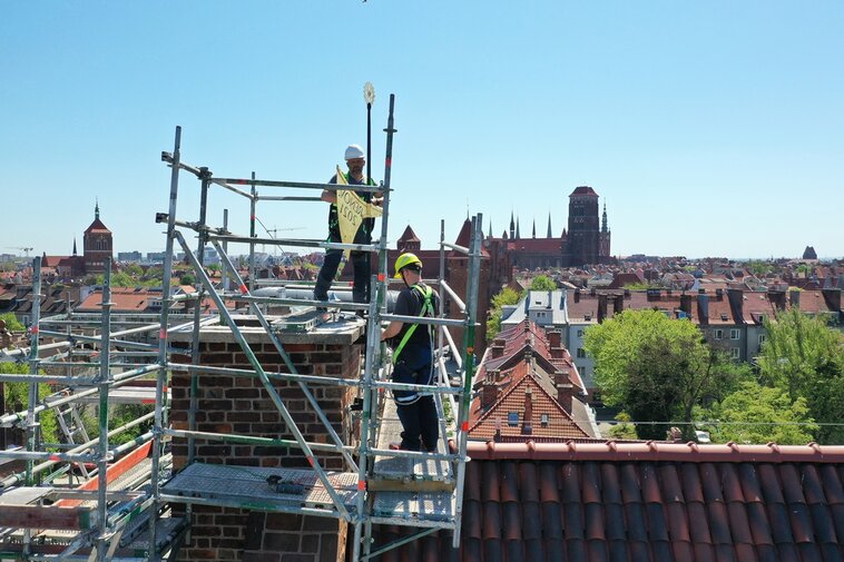 Dwaj mężczyźni stoją na rusztowaniu wokół komina Wielkiego Młyna. Montują złotą chorągiew wiatrową o kształcie topora. Widać napis Renov. 2021.