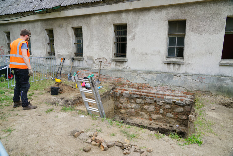 Zdjęcie. Przestrzeń. Budynek z niewielkimi zakratowanymi oknami, pokryty szarym tynkiem. Poniżej wykop. Archeolog patrzy w wykop i odsłoniony fragment fundamentów koszar napoleońskich. 